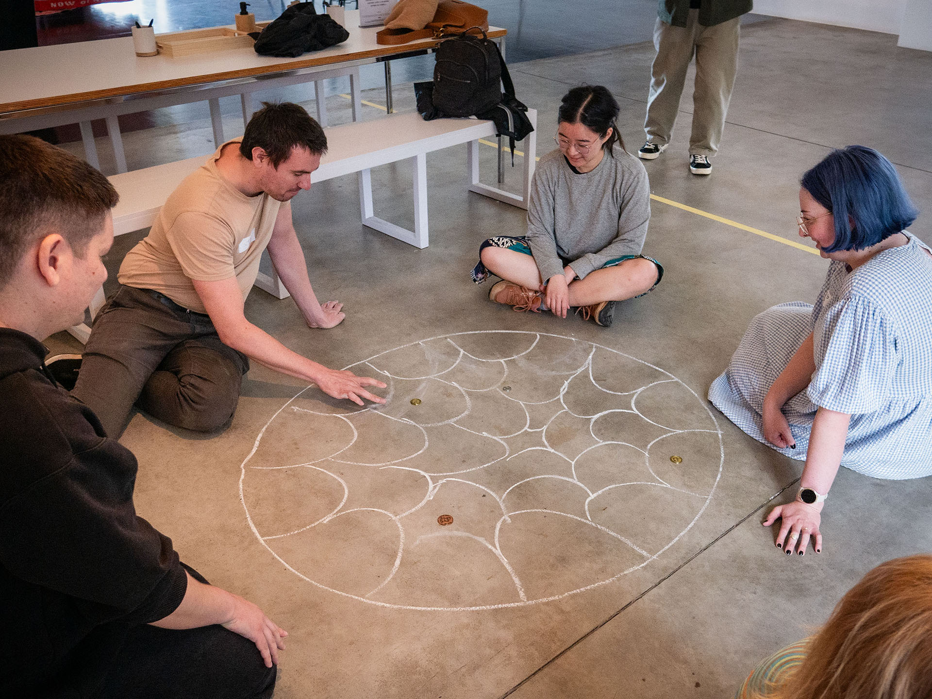 Four participants sitting on the ground flicking a coin for the Traces of Play workshop.